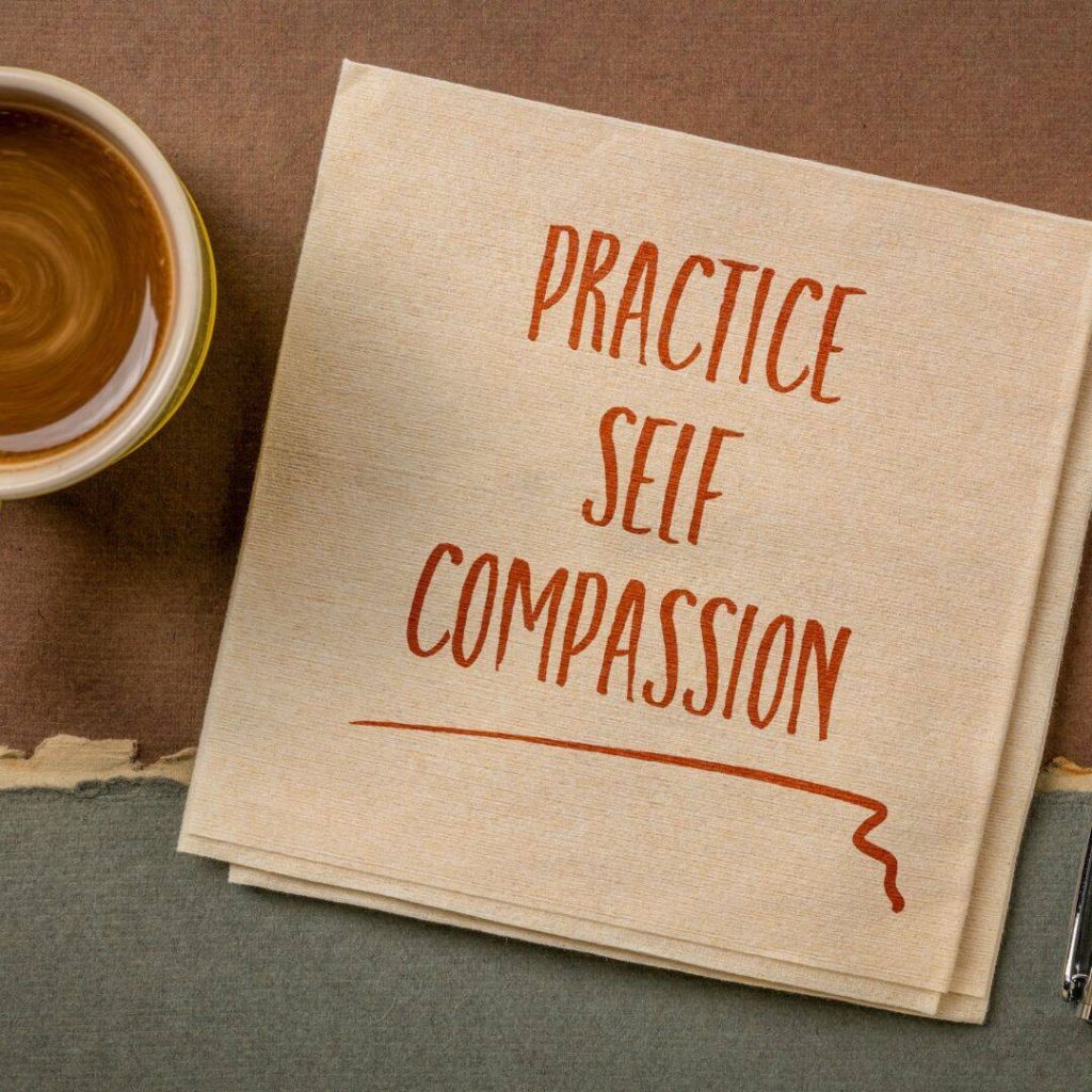 Practice Self-Compassion written on a napkin in orange market with a mug of hot cocoa next to it.