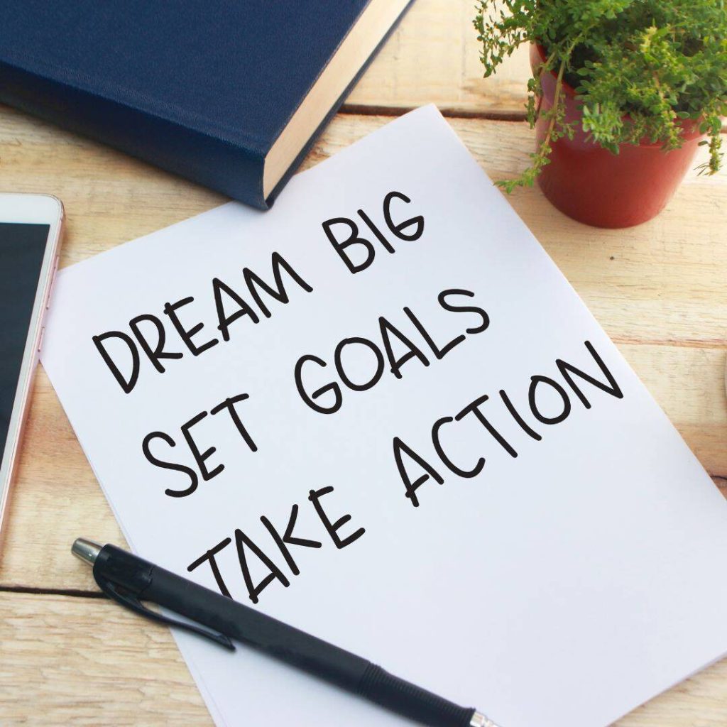 Dream big, set goals, and take action written in black pen on white sheet paper with blue book and phone on table.