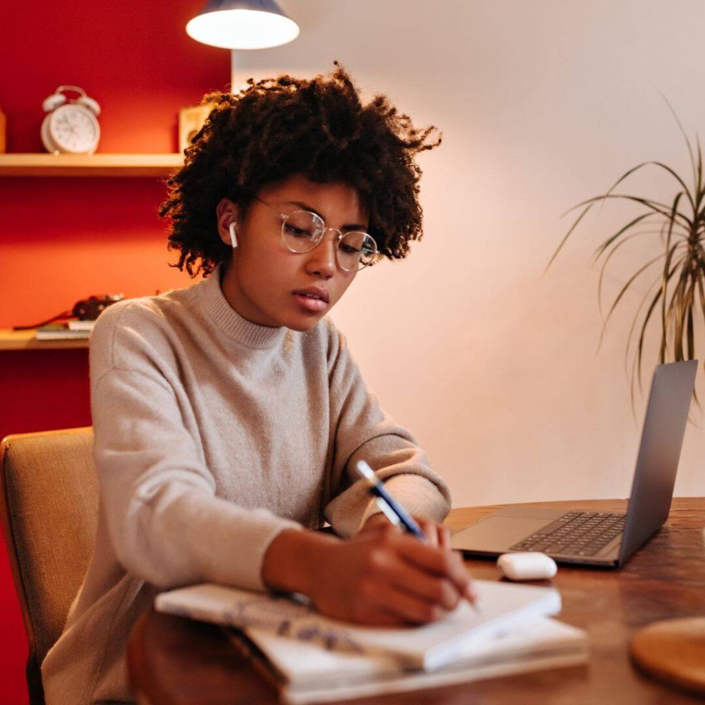 Young woman of color wearing glasses sitting at a table in front of her laptop writing our her intentions in a journal.