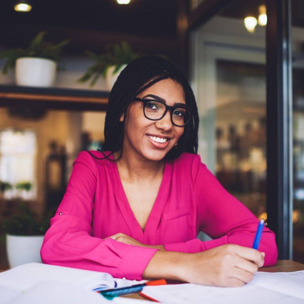 Black woman student wearing glasses, a hot pink blouse sitting at her desk smiling slightly at the camera and holding a blue pen.