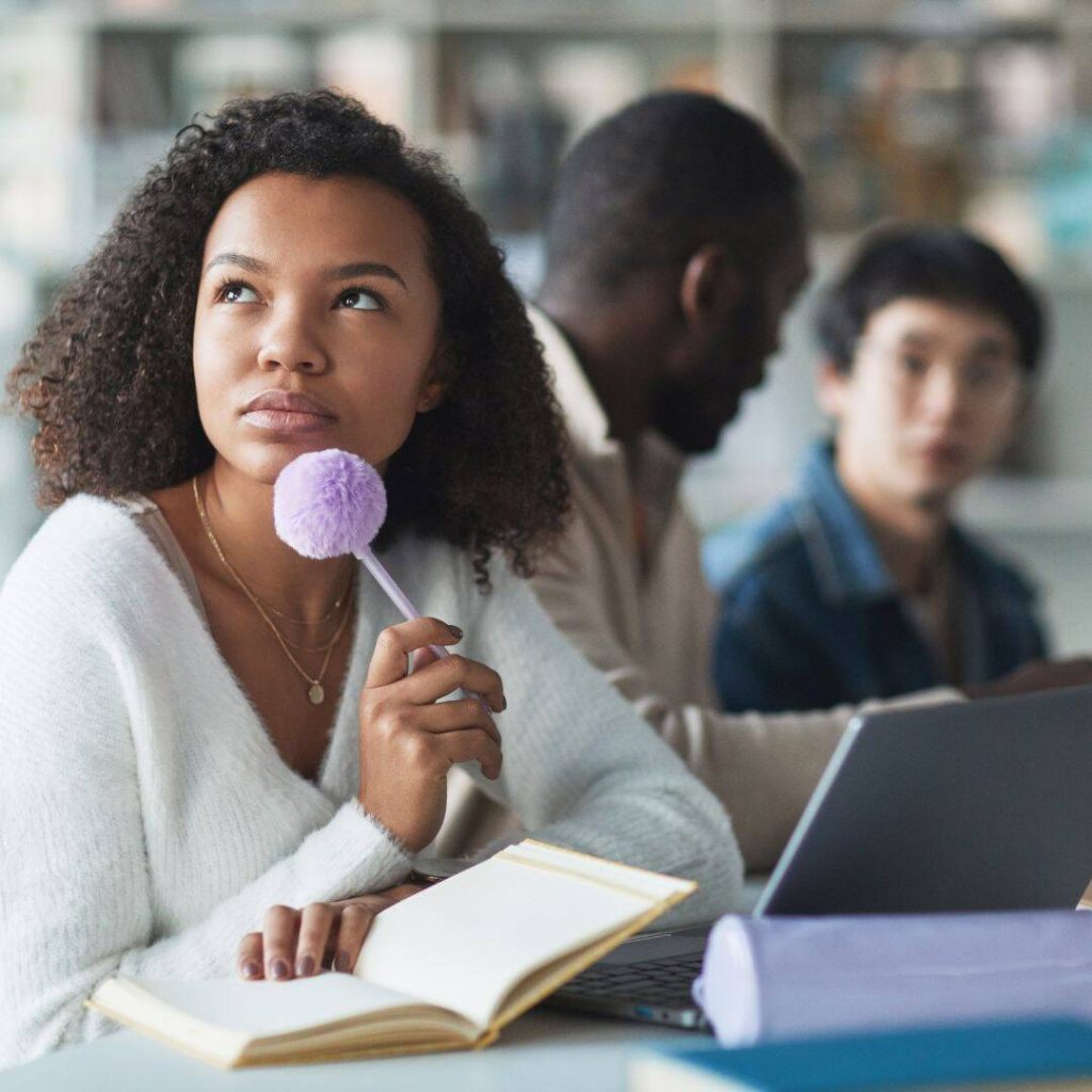 Black woman looking up to the left thinking holding a purple pom pom ball pen in her hand with a book open on the table. 
