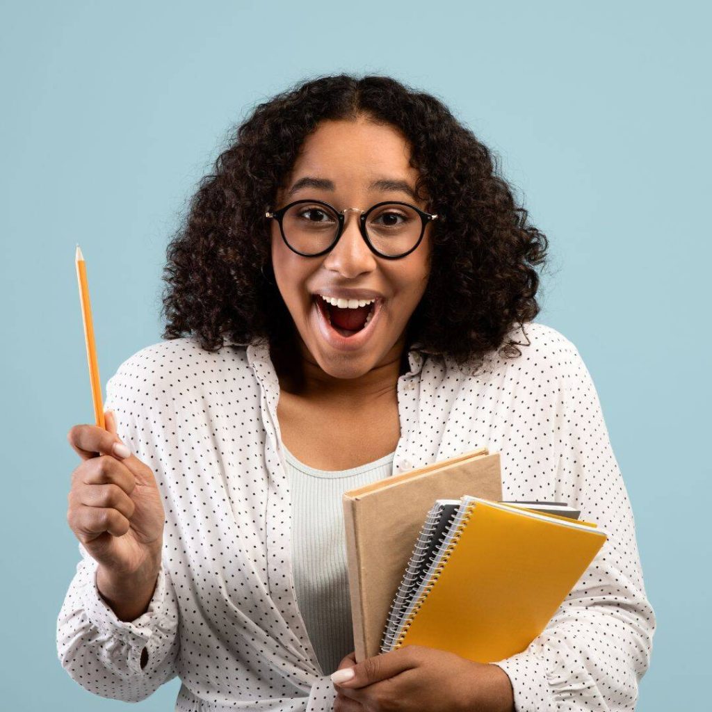 Black woman wearing glasses holding a pencil straight up and having aha moment.  Notebooks in the other hand.
