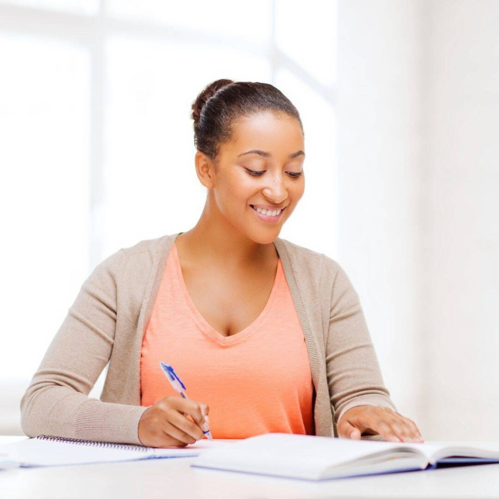 Black woman looking entertained writing a book.  Smiling and holding a blue pen in her hand.