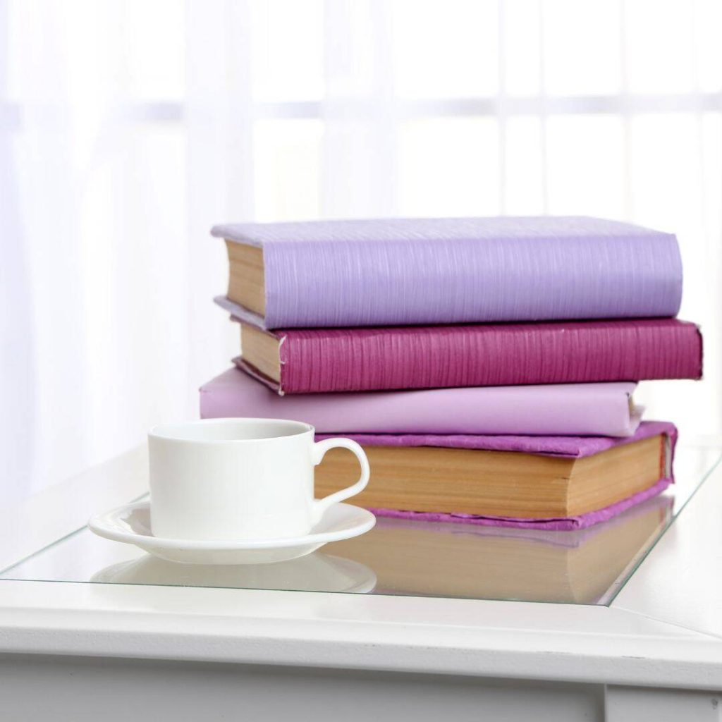 A pile of pink and purple hardback books stacked up on a table with a white mug sitting beside it.
