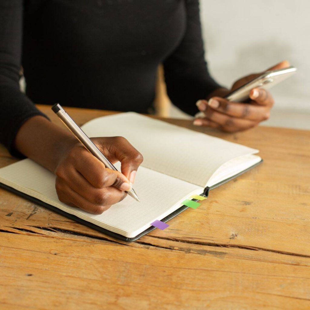 black woman with a black shirt sitting at a wooden desk writing in an open notebook. Holding a cell phone in the other hand.  Just her body is shown, no face. 