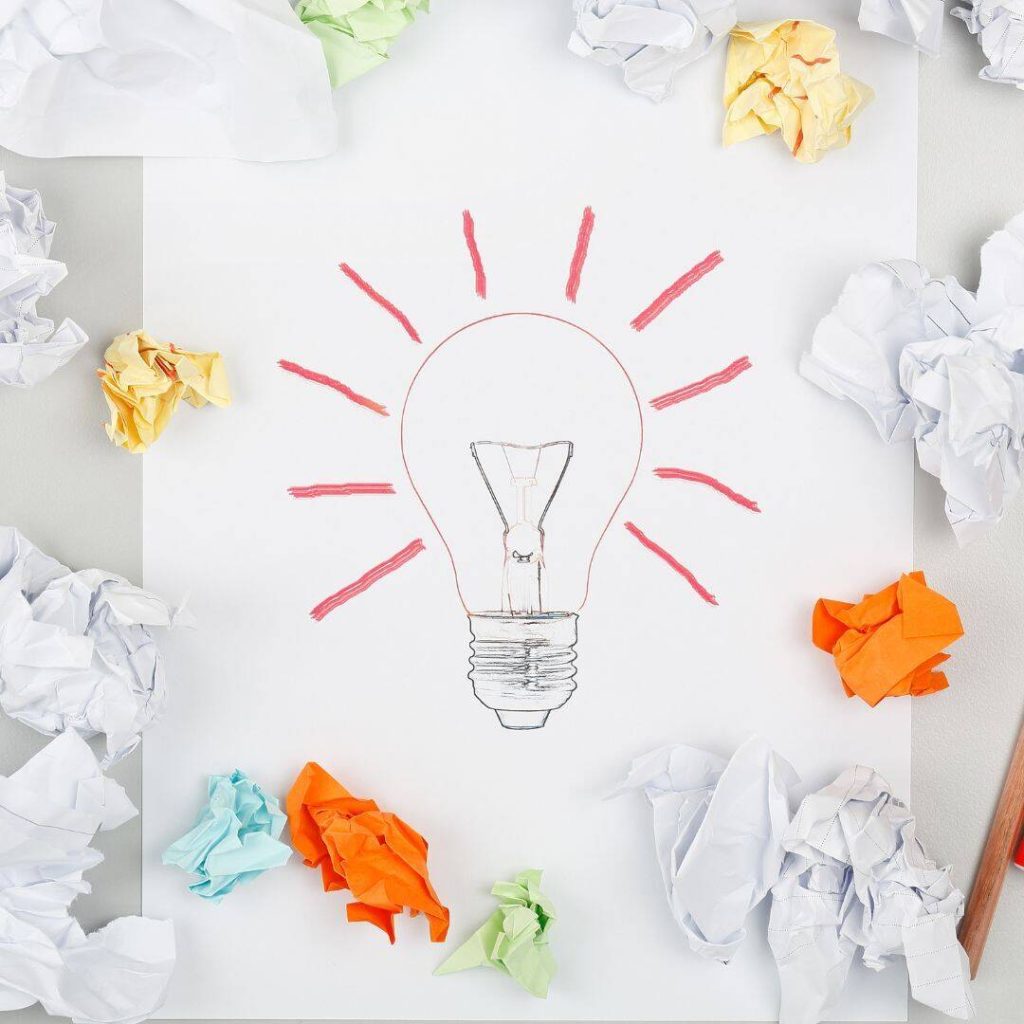 Light bulb drawn on a piece of paper with red lines radiating from it to signify brainstorming ideas.  Balled-up pieces of white paper, bright orange, and blue paper surround the light bulb to symbolize ideas thrown out or scrapped. 