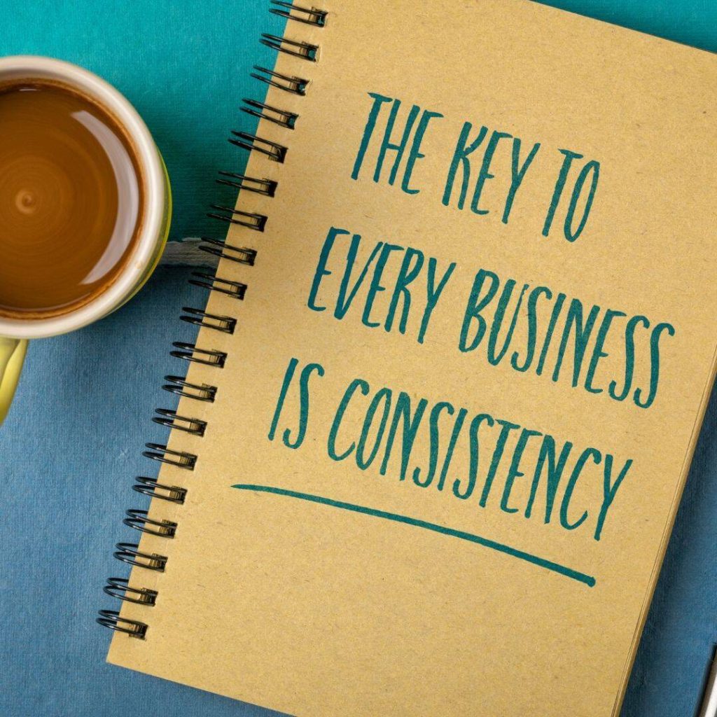 The key to every business is consistency written in Teal on the back side of a spiral bind notebook.  Notebook placed on table with teal table cloth and a mug of coffee sits beside it. 