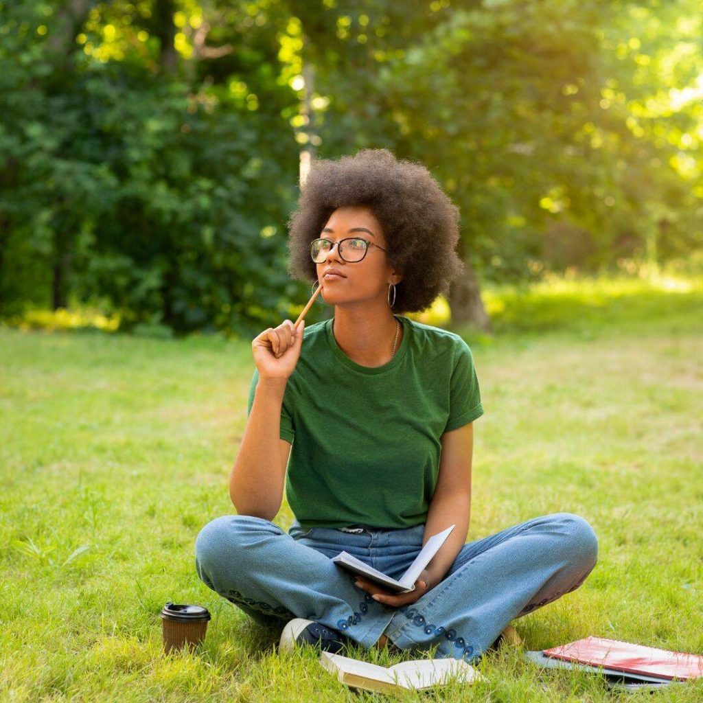 Black woman wearing afro, glasses, and kelly green shirt sitting in the grass with her legs folded.  She is looking up towards the sky, pencil in hand thinking.  She is taking her journaling to the next level by creating a ritual of journaling outside in the park.