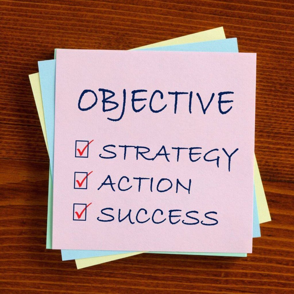 Objective, Strategy, action, and success are written on a light pink post-it. Light blue and neon yellow post-its lying behind it.  All post-its are laid out on a wooden table. 