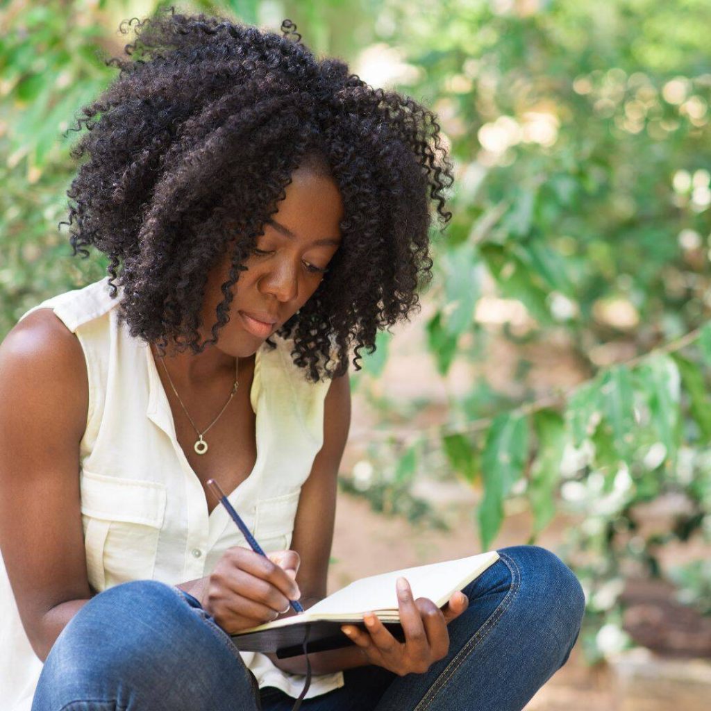 Black woman sitting outside in the grass with her legs folded and journal in her lap.  She's writing and looks as if she's processing her emotions.