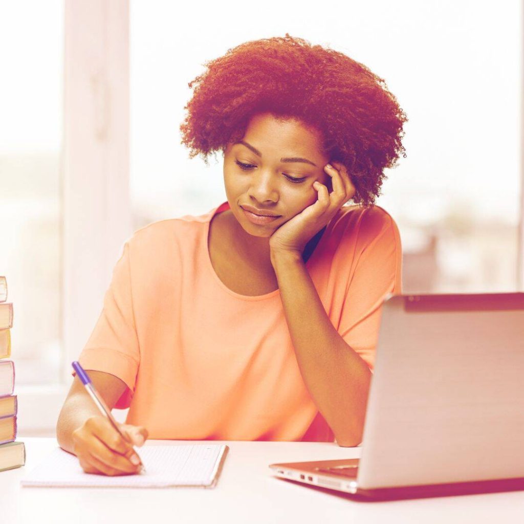 Black woman sitting at her desk journaling looking down.  She looks sad and scared.  There is an open laptop in front of her.  She has her elbow resting on the table, cheek in palm.