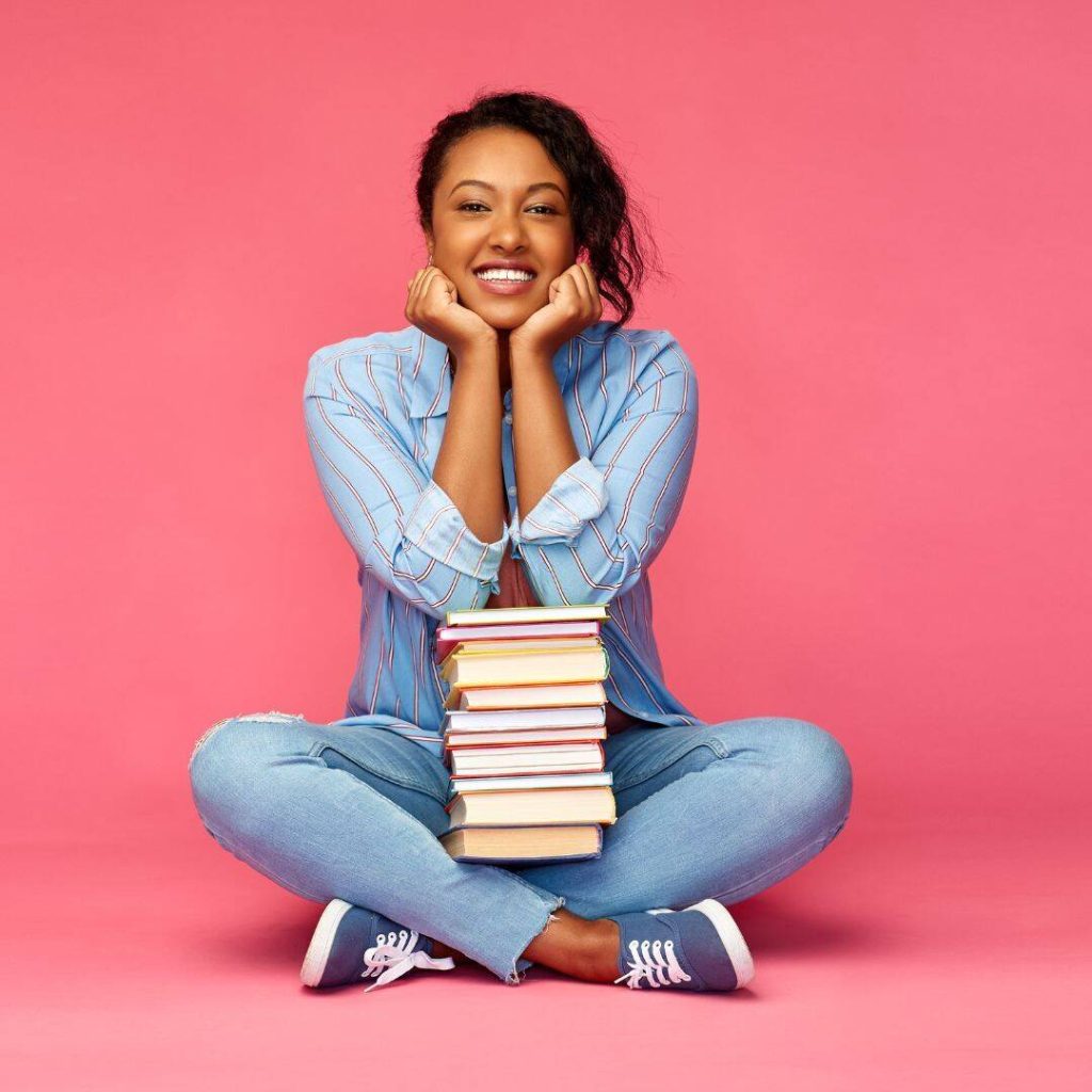 A black woman sitting on the floor, with her legs folded pretzel style.  She has a pile of books in her lap, palms of her hands underneath her chin. She is smiling and wearing a light blue button-up shirt with light blue denim jeans.  Book idea