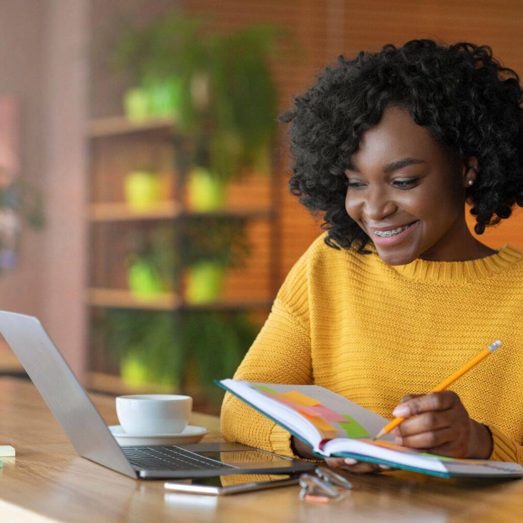 Black woman smiling wearing a yellow sweater staring at her laptop.  Holding open notebook writing in it and looking at 5 tips to start your book manuscript for aspiring writers. 