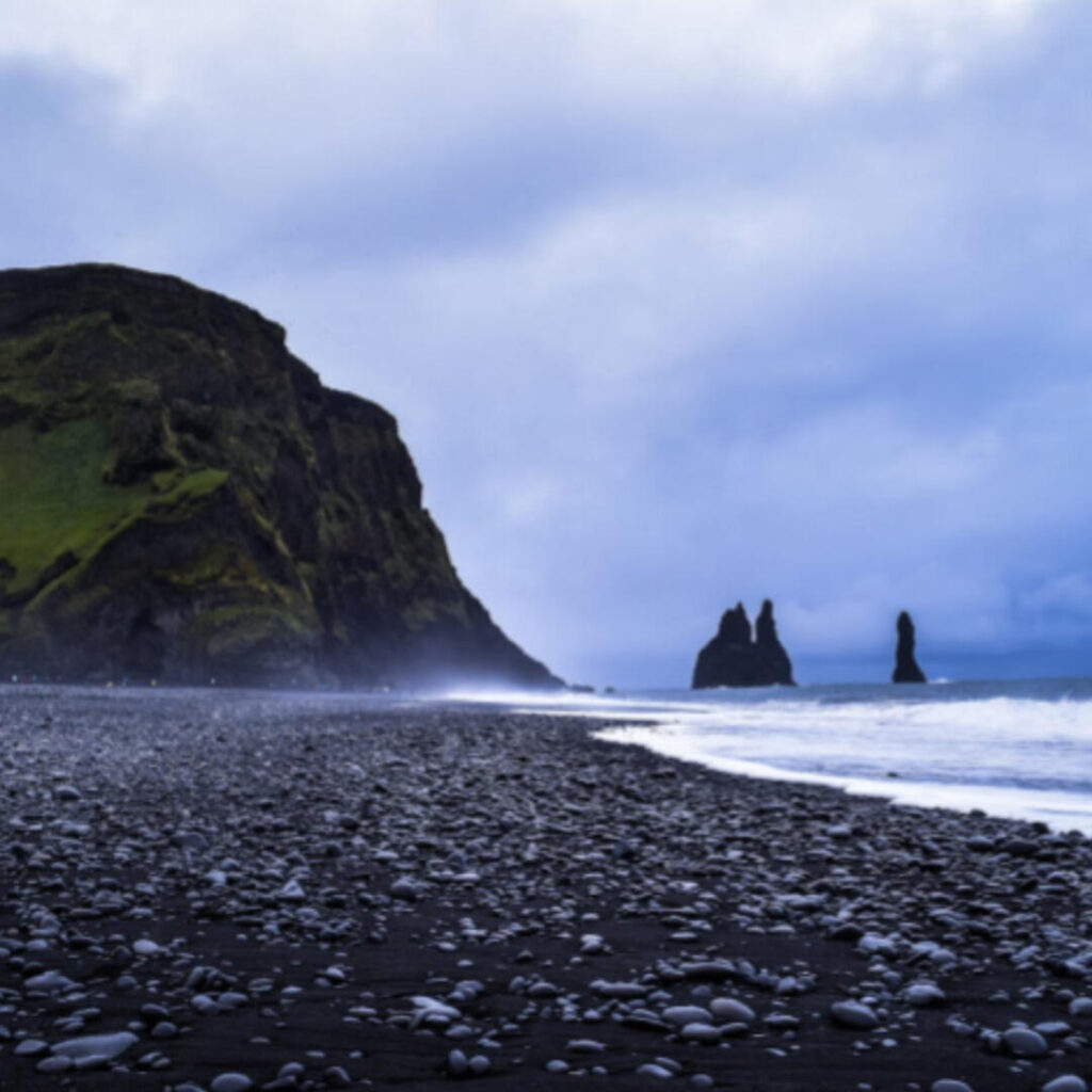 A rocky, black sand beach. A green and brown cliff sits at the edge of an ocean, below blue storm clouds.