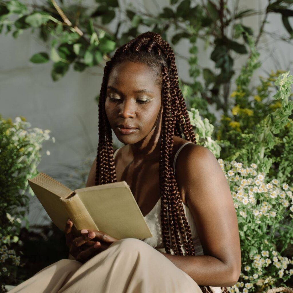 A black woman wearing an olive green dress is siting down reading a book and is surrounded by green plants and small white flowers.