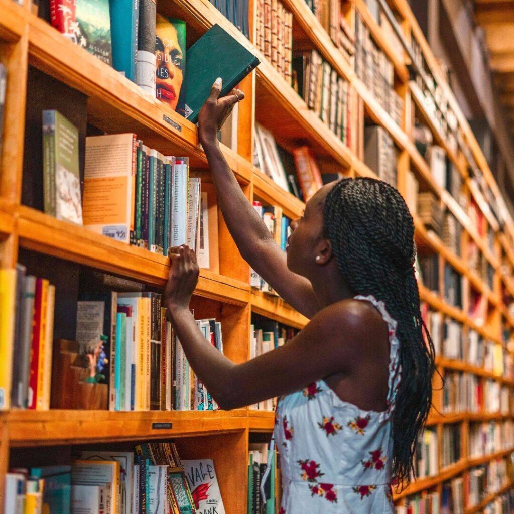 A black woman wearing a light blue top is selecting a book from a shelf in a library.